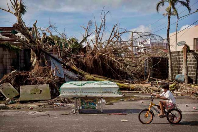TACLOBAN, PHILIPPINES - NOVEMBER 13: A young boy cycles past a coffin amid scenes of devastation in the aftermath of Typhoon Haiyan on November 13, 2013 in Tacloban, Leyte, Philippines. Typhoon Haiyan, packing maximum sustained winds of 195 mph (315 kph), slammed into the southern Philippines and left a trail of destruction in multiple provinces, forcing hundreds of thousands to evacuate and making travel by air and land to hard-hit provinces difficult. Around 10,000 people are feared dead in the strongest typhoon to hit the Philippines this year. (Photo by Kevin Frayer/Getty Images)