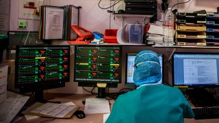 En la foto, un médico con vestuario protectivo sentado enfrente de pantallas.