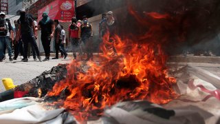 Fogata en la que se quema propaganda electoral; a lo lejos se ve a un grupo de jóvenes que protestan