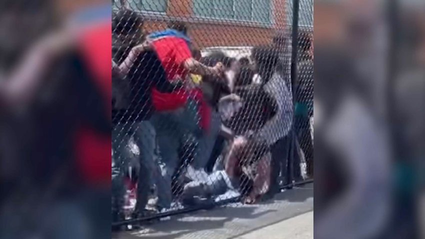 A group of teens is seen beating a grounded teen who is up against a chain-link fence.