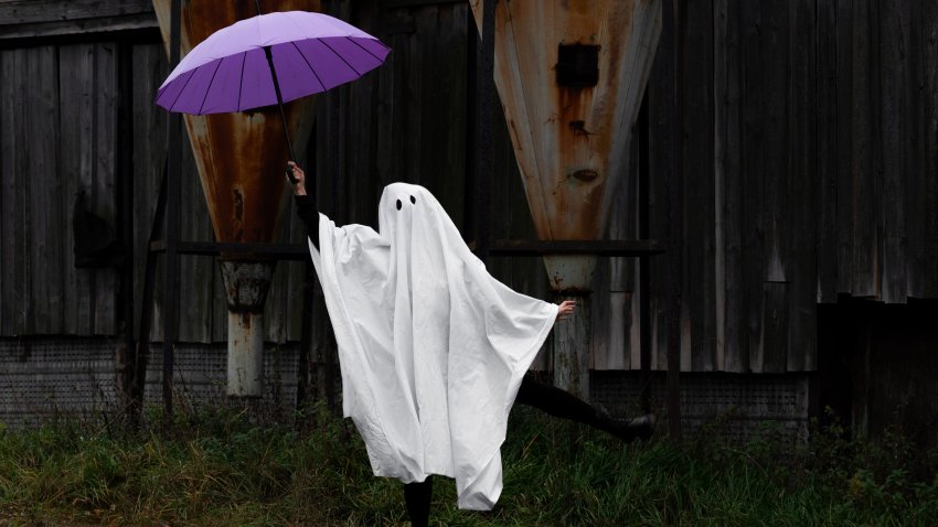 Ghost woman in a white sheet holds a purple umbrella and dances near an old wooden building with rusty metal hoods. Halloween concept.